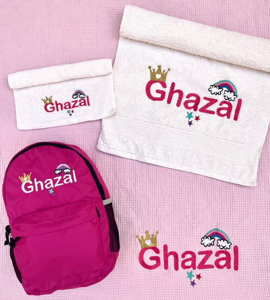 A set of personalized nursery items including a pink backpack, a white blanket, and two white towels with embroidered text and decorative elements in different colors.