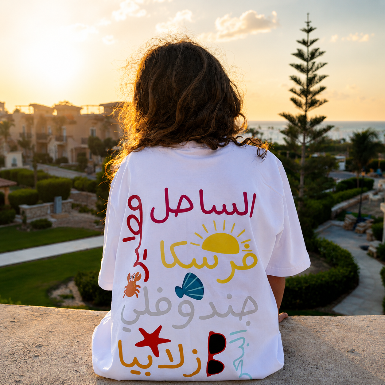 Person wearing a white t-shirt with colorful designs and Arabic text, standing on a balcony with a scenic view.